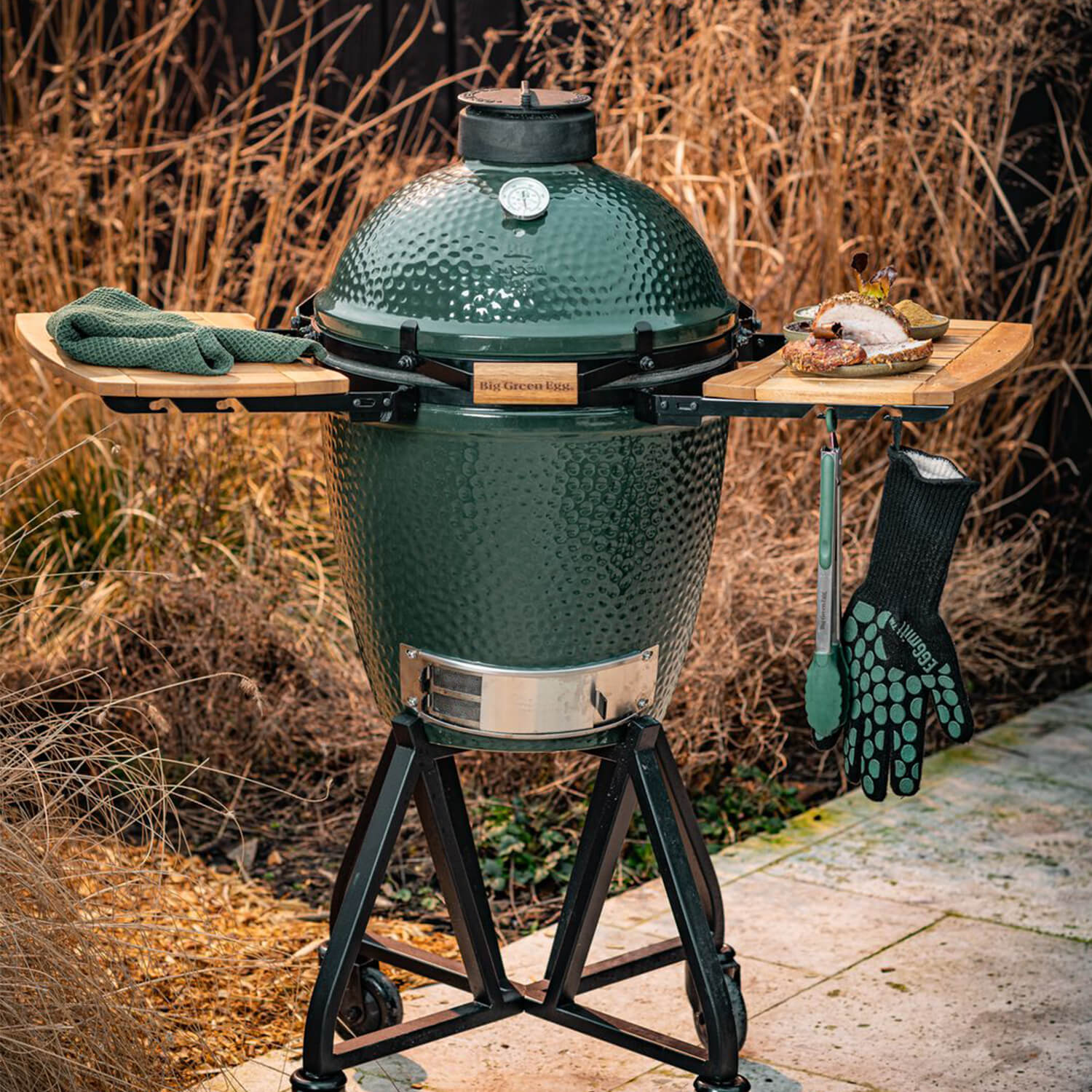 Medium sized big green egg with multilpe accessories including eggmitt, tongs, side shelves and sitting on the egg nest.